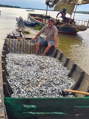 Zeb Hogan sits with a boatload of fish in a fishing boat on the Mekong river with other boats around him