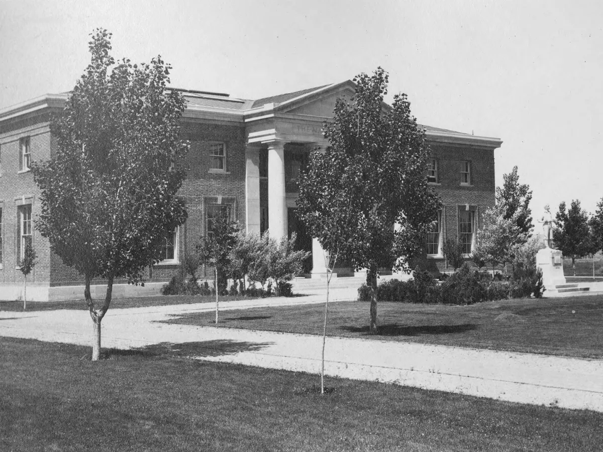 Black and white photo from 1908 of two men walking past newly-planted trees near the Mackay School of Mines Building and the Quad. 