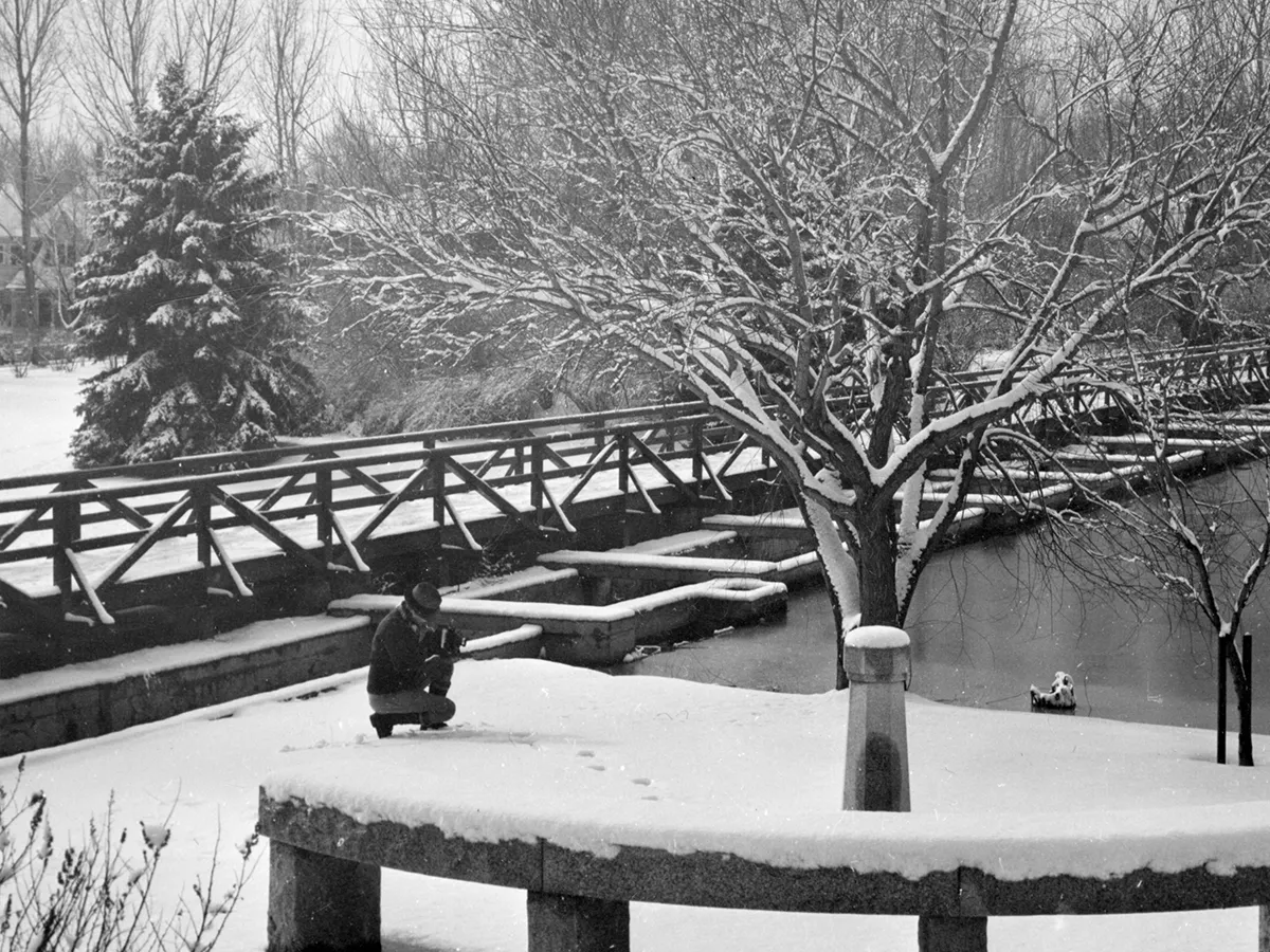 1944 black and white photo of a man kneeling down with a camera next to a snow-covered Tram with Manzanita Lake in the background.