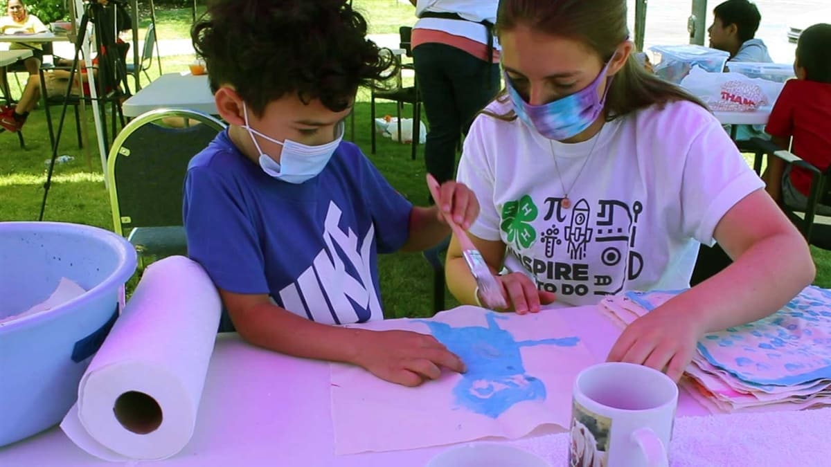 A 4-H youth, who is wearing a mask, is outside on a sunny day. He is sitting under a shade canopy at a folding table, holding a paintbrush. There is a roll of paper towels and a cup of paint-muddled water nearby. He's with a masked 4-H leader. They're working on a STEM project.