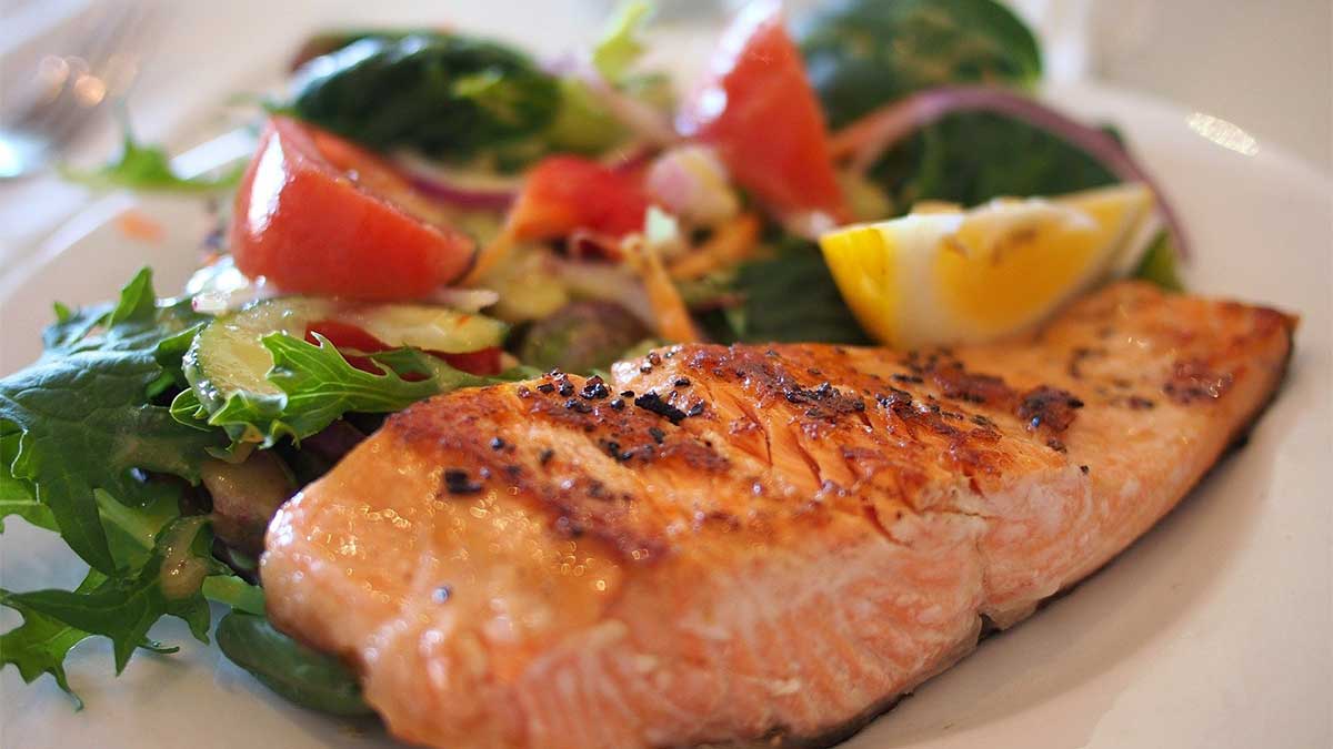 On a white plate is a freshly-prepared meal of seasoned, pan-seared salmon and a salad of leafy greens topped with cucumbers, tomatoes and onions. The dish is garnished with a lemon wedge.