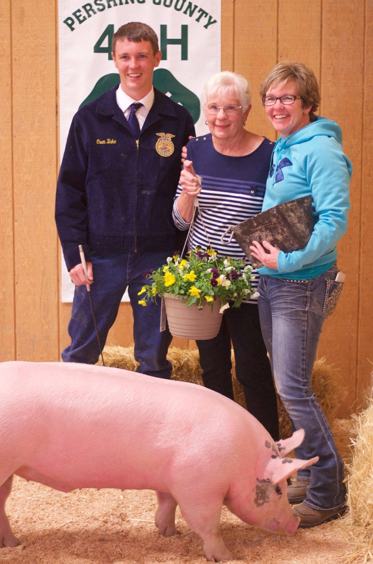 Owen Bake, Sharon Montrose and Shauna Bake stand together in front of a Pershing County 4-H banner and hay bale next to an impressive pink pig.