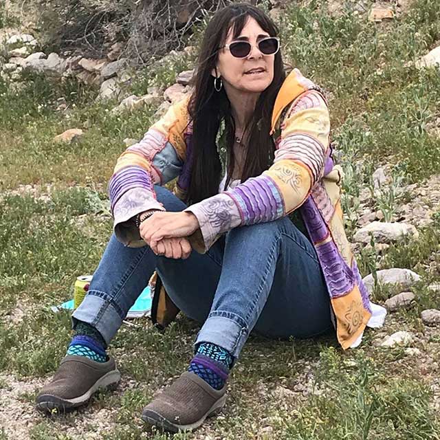 Robin Flinchum wearing sunglasses and a brightly colored sweater, sitting on the ground, arms around her knees