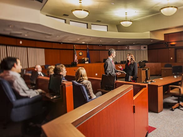 Two people are talking before a jury box full of people in a courtroom. 