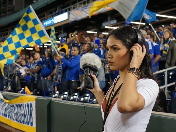 A student holds a microphone up to a crowd in a soccer stadium while listening in headphones.
