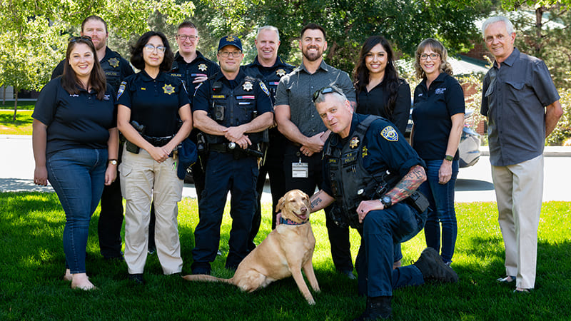 Service dog and police officers in uniform standing on the grass under the shade of trees on campus in front of the Fitzgerald student services building