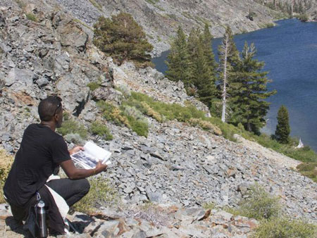 Student doing field research sitting on a steep, rocky slope looking down at a mountain lake and holding a notepad