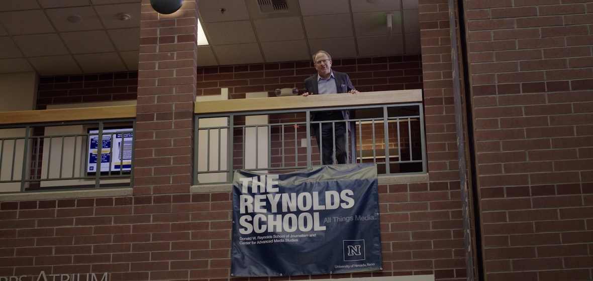 Man stands on the second floor of an atrium with a sign that reads The Reynolds School below him.