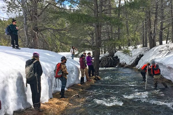 A professor leads a team of students researchers through a stream surrounded by snow