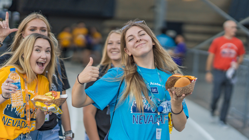 A group of students holding food and drinks give a thumbs-up to the camera during NevadaFIT.
