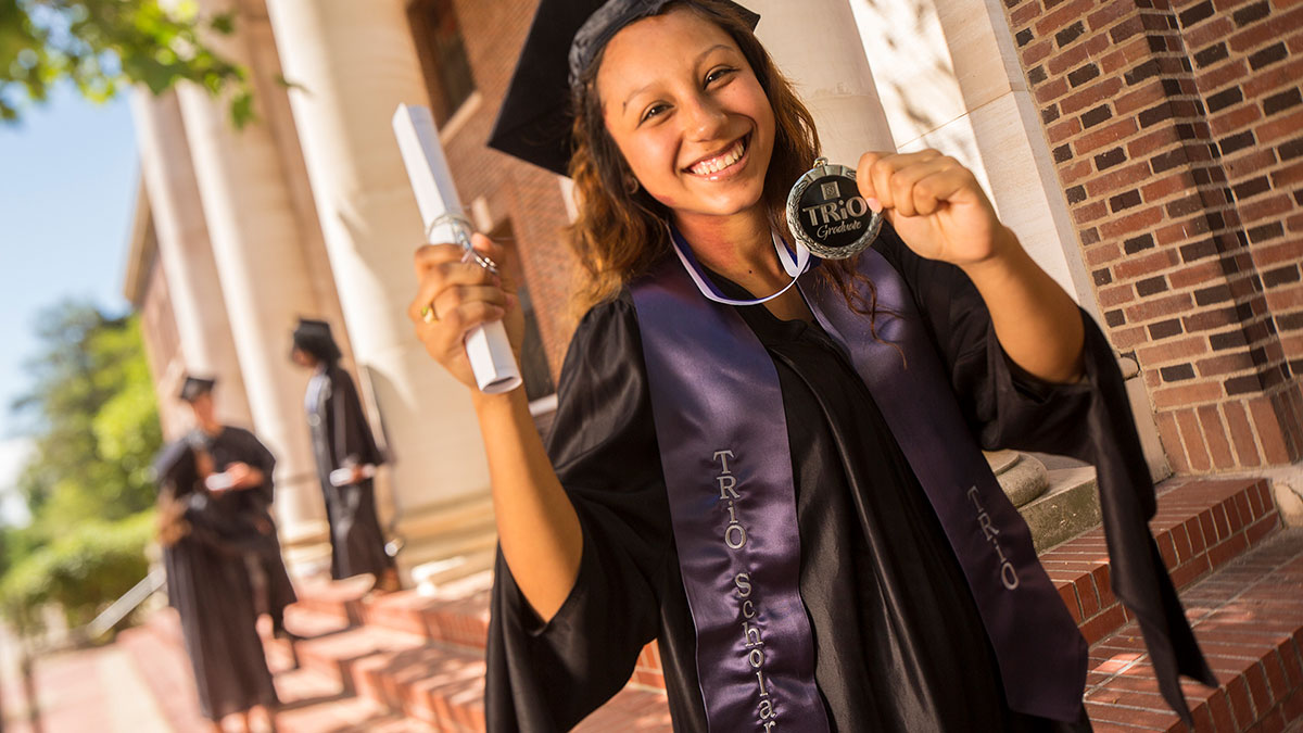 A Trio graudate wearing a cap and gown and holding up a Trio Graduate medal