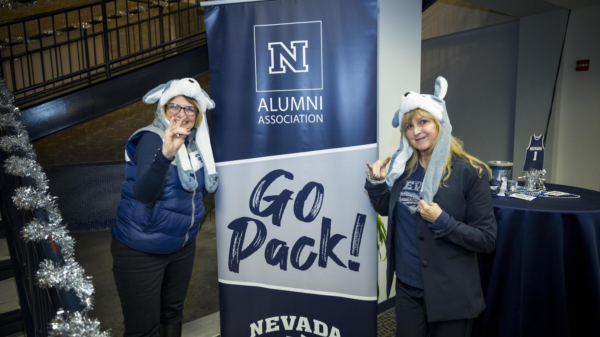 Two alumnae posing next to a Go Pack! sign wearing wolf pack gear.