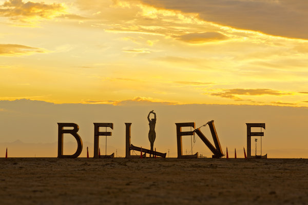 BELIEVE sculpture, which is the word spelled out in large, rusty iron lettering, at Burning Man on the Playa at sunset with large scultpure of a woman in the background