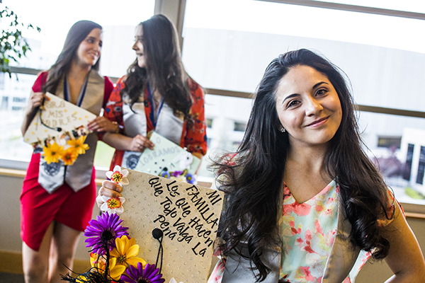 Graduates show off decorated caps outside the Latinx Graduate Celebration, with the cap in the foreground reading "Mi familia, los que hacen que todo valga la pena"