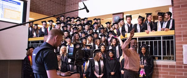 Students and faculty pose in regalia indoors while listening to Paul Mitchell.