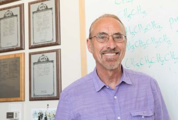 Professor stands in his office; plaques are on the wall behind.