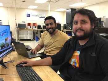 2 men look at camera while seated at table in front of computers