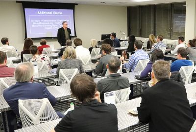 Man is standing and addressing a room full of people sitting at tables.