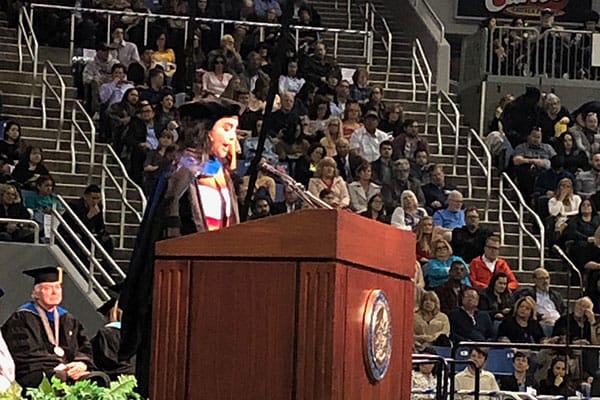 A woman, wearing regalia, speaks to a crowd indoors.