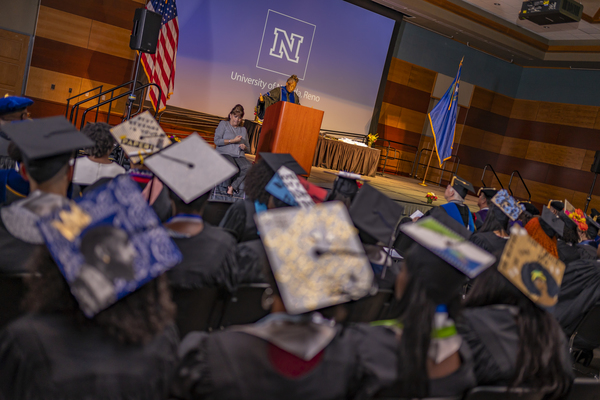 A group of mortar boards, some decorated, on the haeds of graduates with a speaker addressing them in the background at the African Diapspora Graduate Celebration