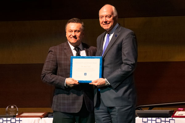 A faculty member and the university president pose on stage indoors.