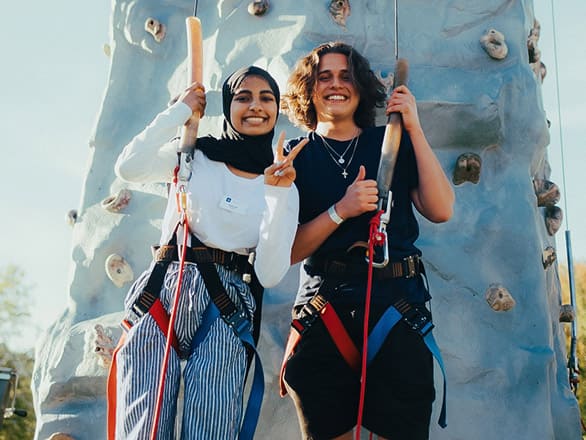 Two individuals stand in front of a rock climbing wall, wearing harnesses and holding climbing ropes, smiling and ready for an outdoor climbing activity.