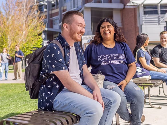 Two people sit on a bench outdoors, smiling and chatting. One wears a "Nevada Wolf Pack" t-shirt. The background includes other people, buildings, and trees.