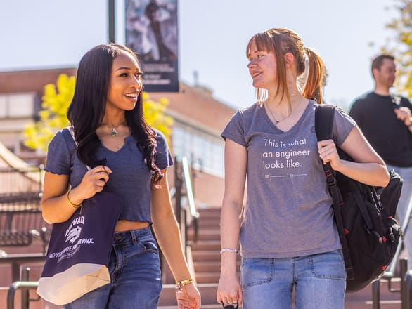 Two individuals walk outdoors with backpacks, one holding a "Nevada Wolf Pack" tote bag and the other wearing a shirt that says "This is what an engineer looks like." They are smiling and talking.