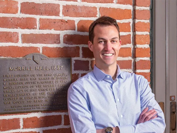 Ethan Ris poses with his arms folded in front of Morrill Hall, next to the hall's dedication plaque.