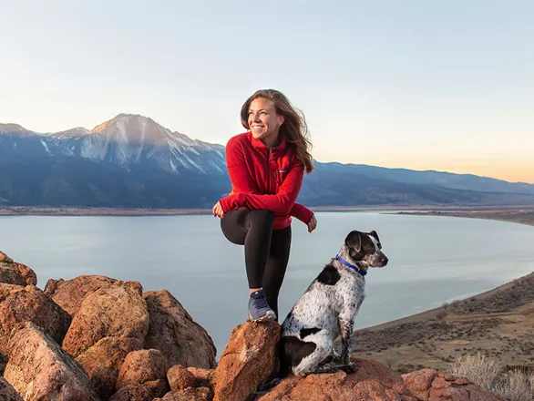 Krishna Pagilla stands next to her dog and bike on a cliff overlooking a lake in the valley behind her.