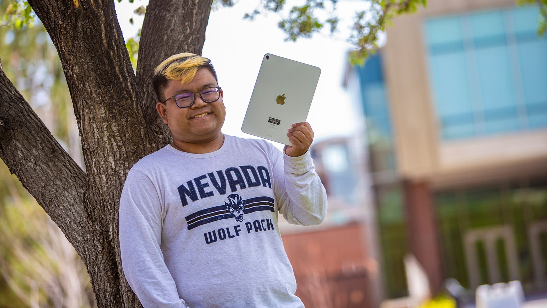A student wearing a shirt with the words "Nevada Wolf Pack" on it holds up an iPad while leaning against a tree.