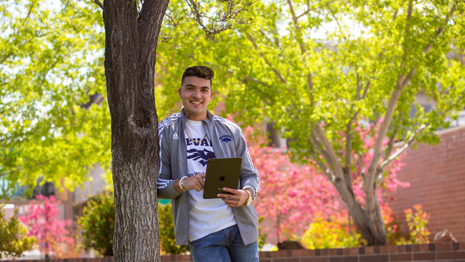 A student leans against a tree while holding a rose-colored iPad.