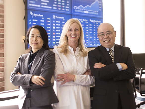 Three Finance faculty members stand in front of a stock ticker display with arms folder.