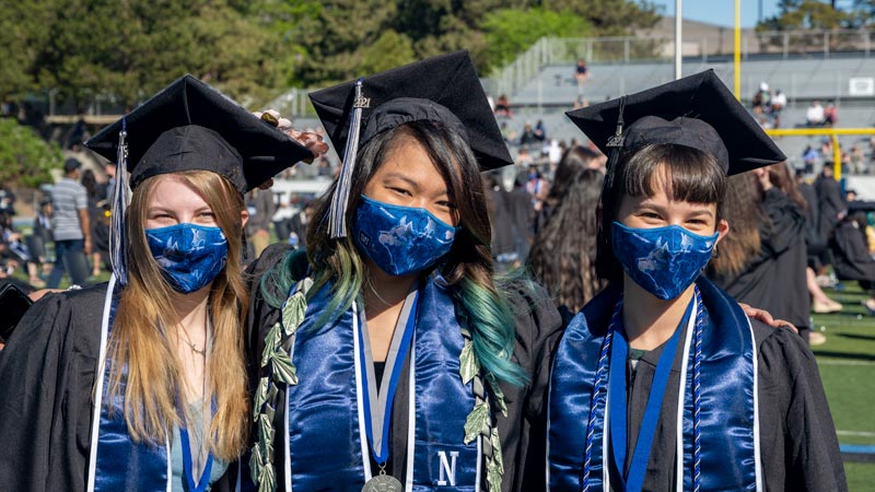 Three people standing arm in arm in caps and gowns on the quad at commencement