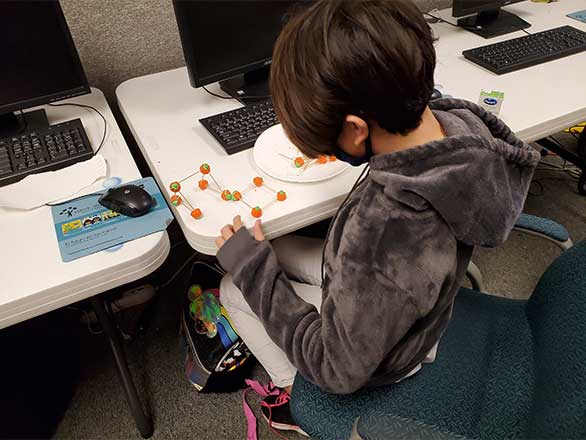 A boy wearing a mask and a comfy hoodie sits at a computer table. He has a paper plate full of toothpicks and green-topped, orange pumpkin-shaped Halloween candy in front of him. Some pieces of candy are connected to others with toothpicks, forming a futuristic 3-D shape.