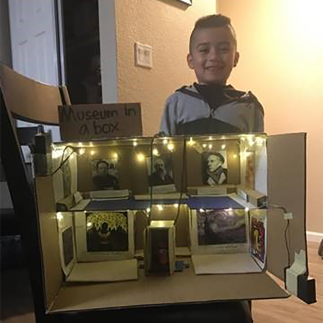 A young child with his family's museum in a box diorama. It's made from cardboard and two stories tall. Famous paintings line the walls, and twinkle lights adorn the spaces.