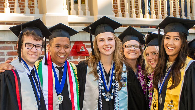 Six people standing arm in arm in caps and gowns on the quad at commencement