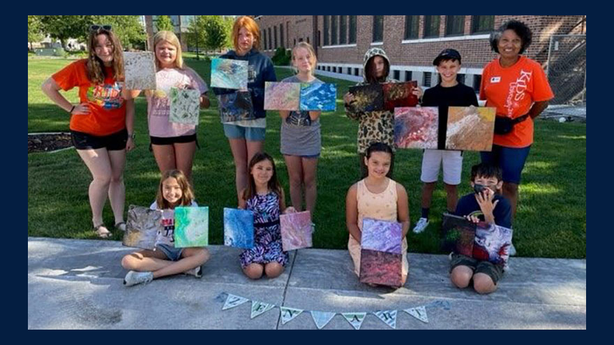 Group of children and two women displaying paintings they made at camp. 