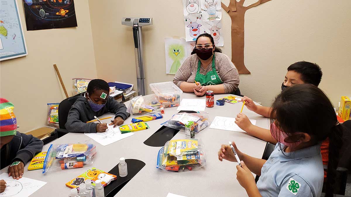 Samantha Shoupe and 4-H youth sit around a long rectangular table covered in craft supplies as they each draw reindeer. Educational posters (including one of the solar system) and kids' artwork adorn the walls.