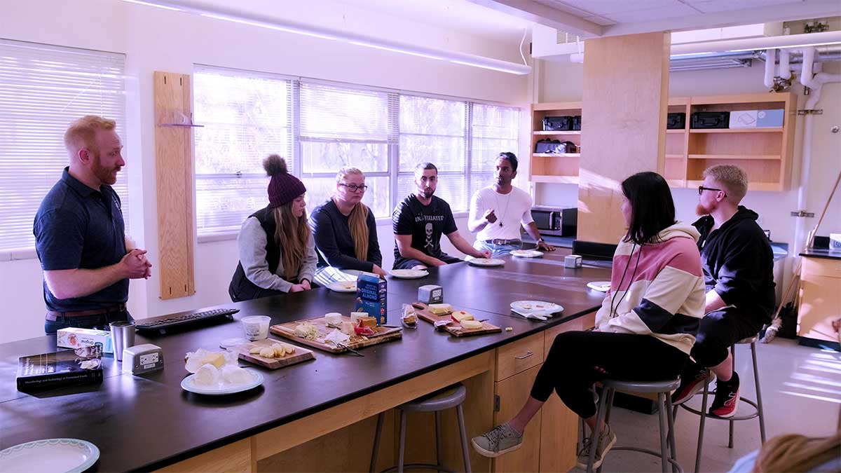 A professor and students around a lab table with water activity of foods supplies, including cheese, fruit and crackers.