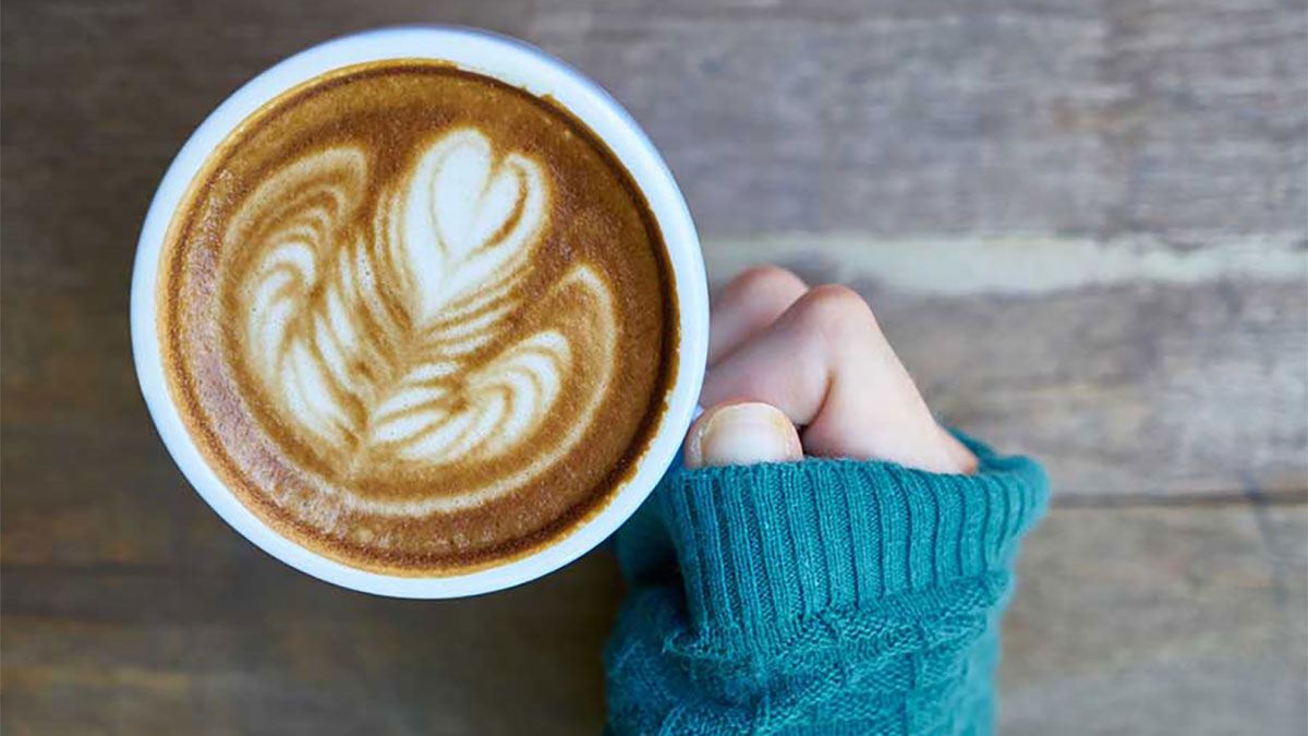 A hand holds a full coffee mug by the handle above a wooden table