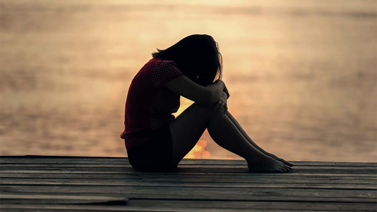 Silhouette of girl on a dock resting her forehead on her knees.