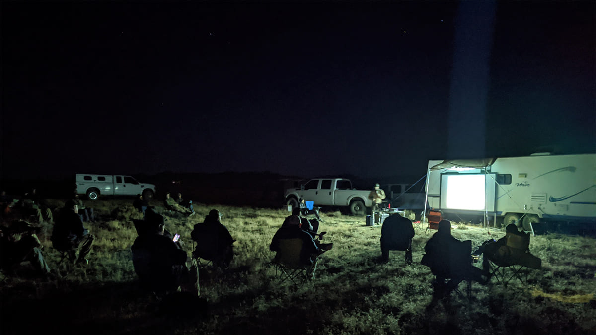 Students sit a safe distance from each other in lawn and camp chairs on a range in the dark, learning from an instructor who stands next to a portable projection screen dramped from an RV.