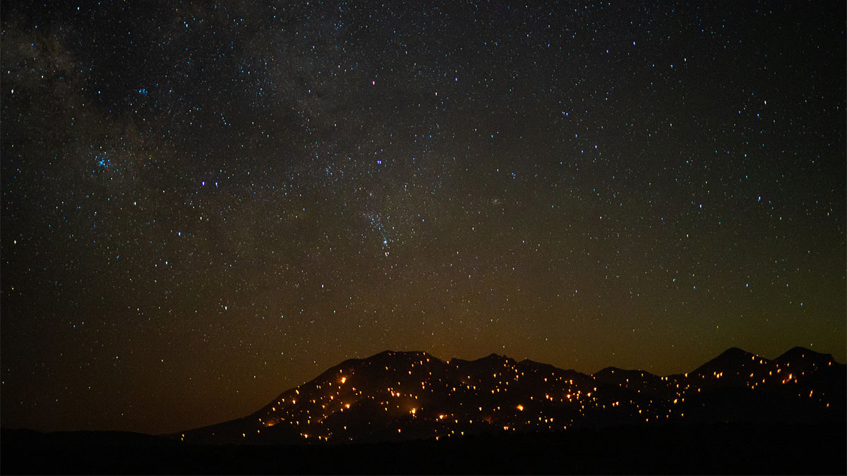 A mountainside on fire at night, it's dark silhouette dotted with flame, mirroring the contrast of dark sky to stars, above