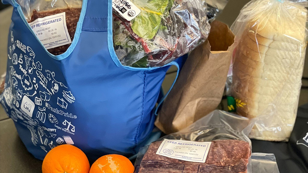 A bag full of fresh ground beef, greens and other food items sits on a table next to a bagged loaf of bread and two clementines and a stack of two packages of ground beef.