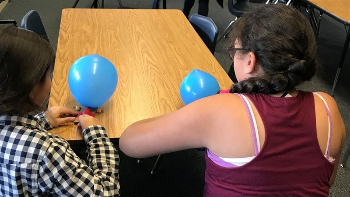 two students use balloons to race cars across a table