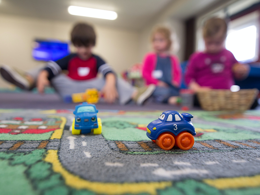 Toy cars on a track in the foreground, in the background there are three children playing who appear blurry.