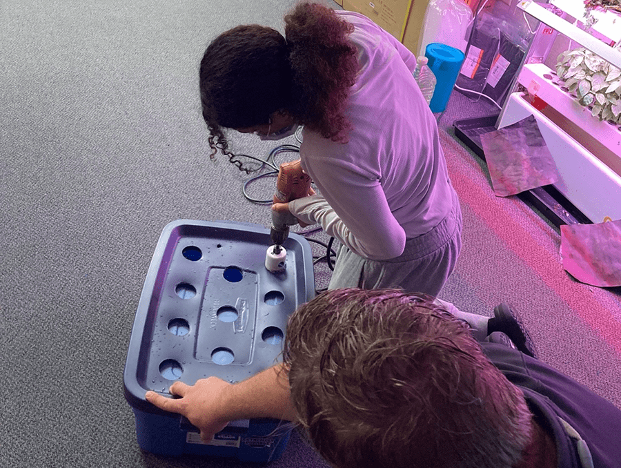 Instructor Taylor Quiram helps a student with a hole saw while creating a hydroponics unit.