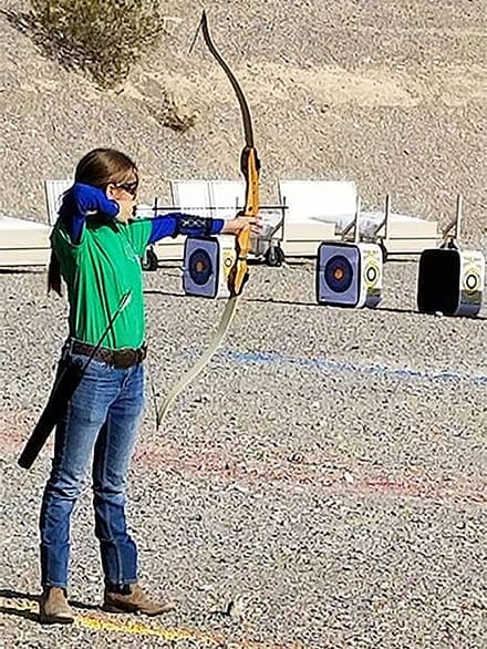 A 4-H'er wearing safety gear on an archery range firing line trains her nocked arrow on a target's bullseye.
