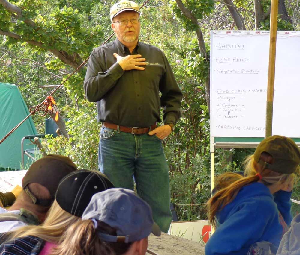 Kent McAdoo, sporting jeans and wearing a ball cap, stands in front of a group of young people. A tent and green trees are in the background. A portable whiteboard with writing on it is to his left.